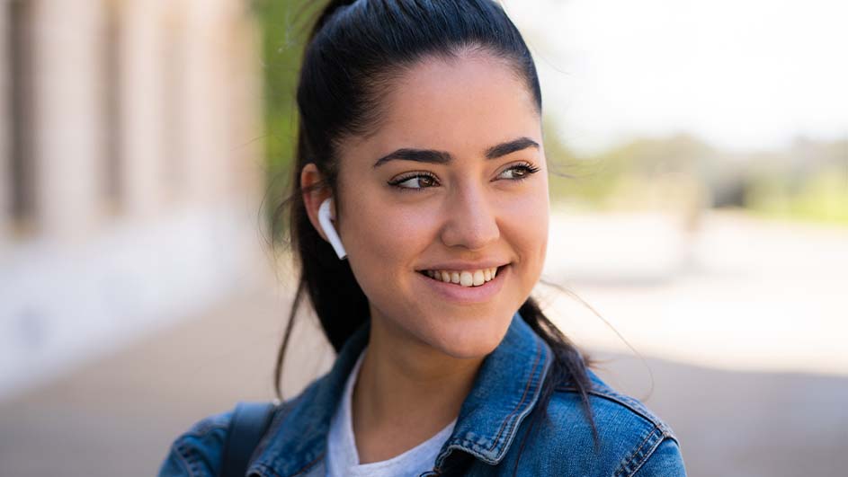 Een lachende vrouw draagt een blauw spijkerjack en witte oortjes buiten, met gebouwen en groen op de achtergrond.
