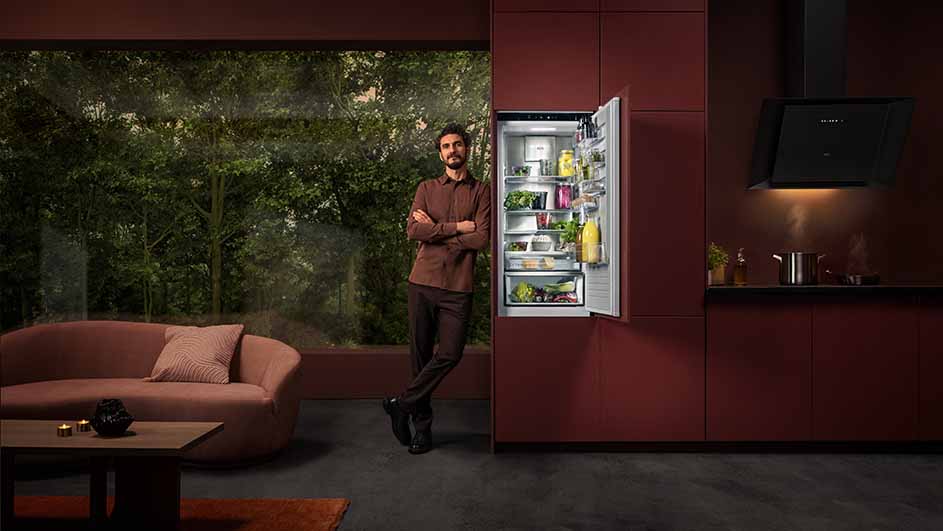 Man leunt tegen een open koelkast in een kamer met een bank, tafel en raam met uitzicht op bomen. Een kookplaat staat rechts.