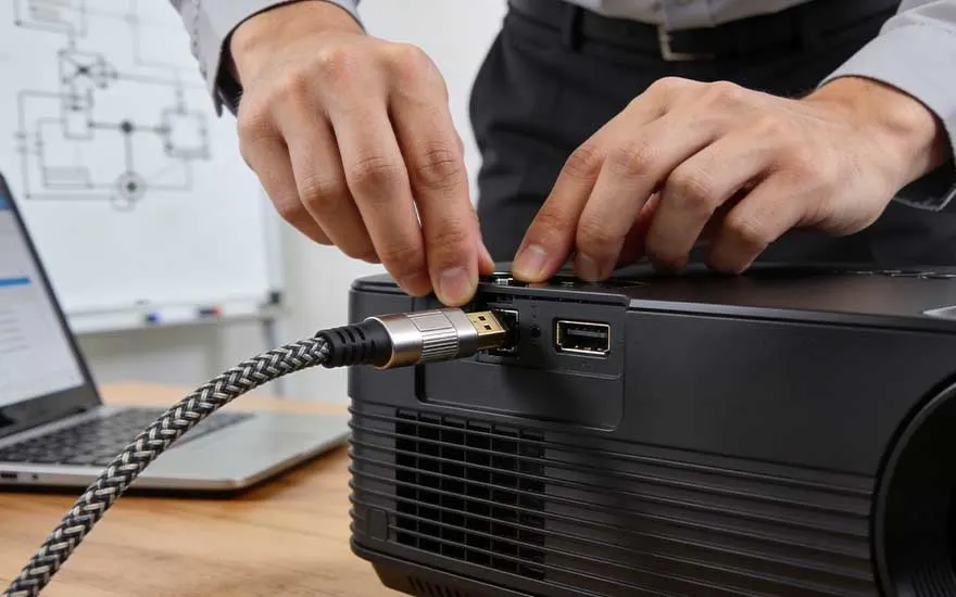 Someone is connecting a cable to a black projector with both hands; an open laptop is also on the table.