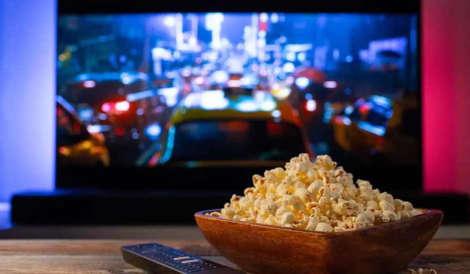Een houten kom met popcorn en een afstandsbediening op tafel. Op de achtergrond staat een tv met een kleurrijke filmscène aan.