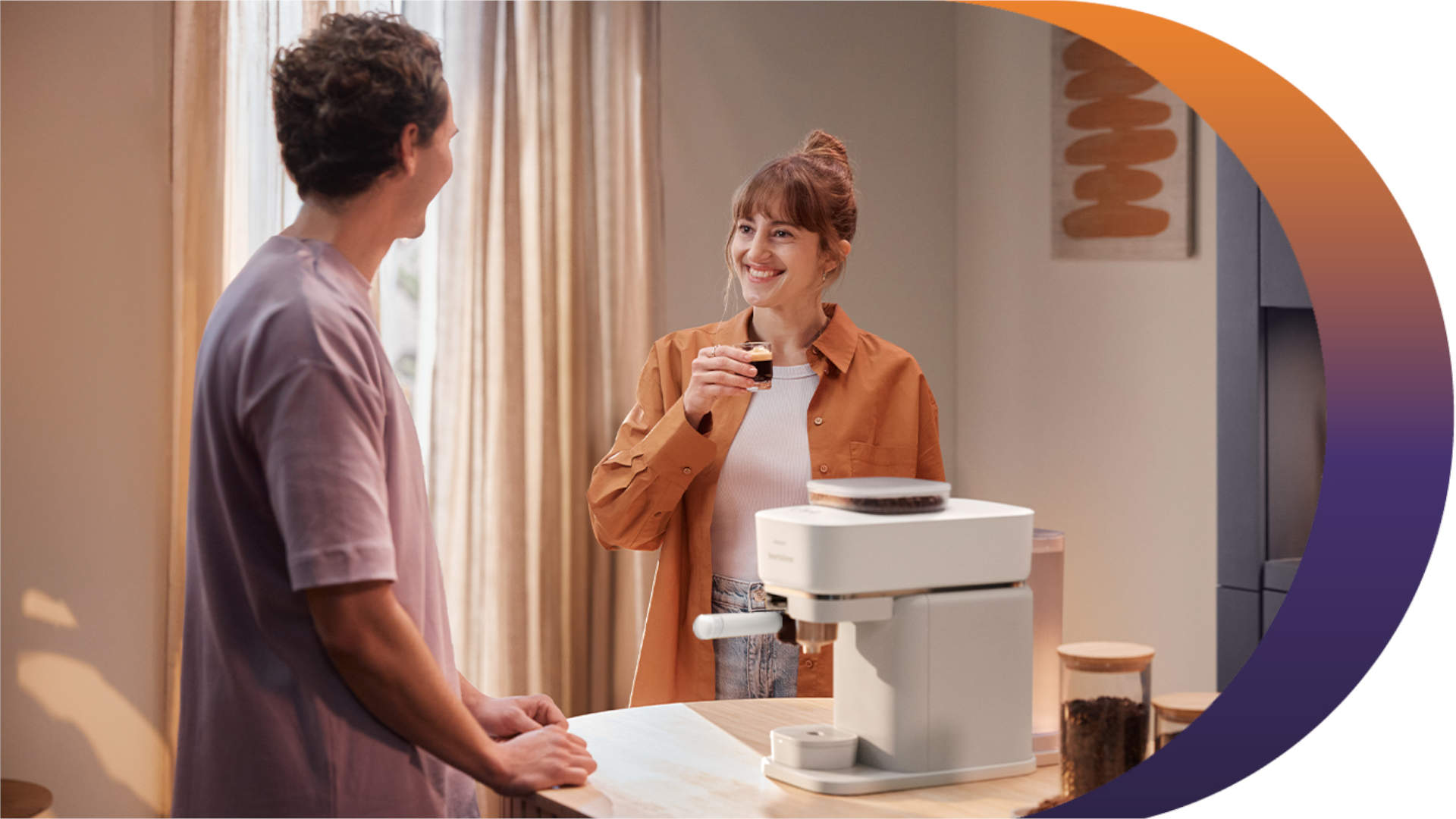 Een vrouw glimlacht met een kopje koffie bij een wit koffiezetapparaat, terwijl een man toekijkt, in een lichte kamer.