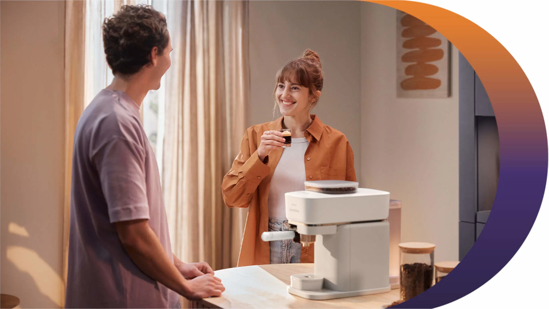 Een vrouw glimlacht met een kopje koffie bij een wit koffiezetapparaat, terwijl een man toekijkt, in een lichte kamer.