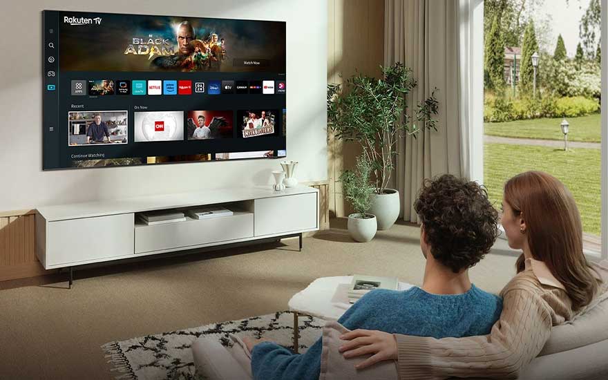 Stel kijkt naar Rakuten TV met Black Adam-film op wandgemonteerde TV in stijlvolle woonkamer met planten en tuinzicht. 