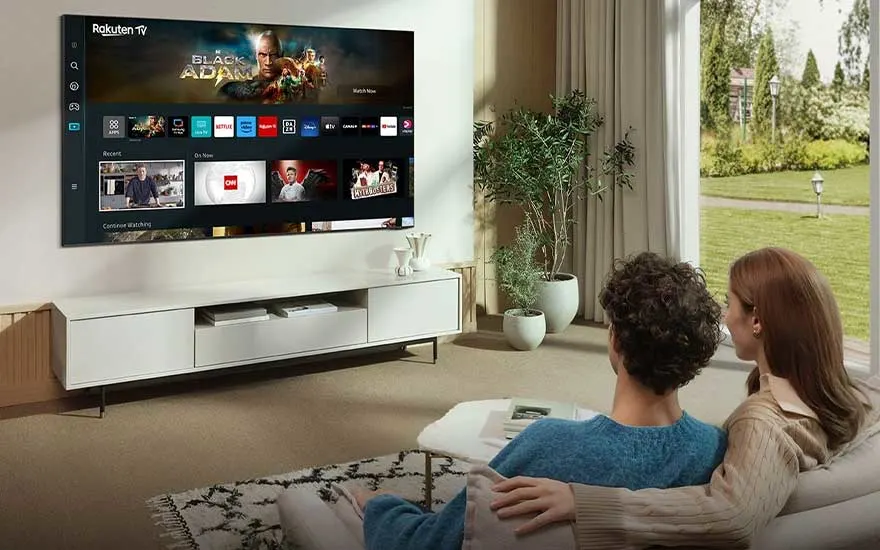 Stel kijkt naar Rakuten TV met Black Adam-film op wandgemonteerde TV in stijlvolle woonkamer met planten en tuinzicht.