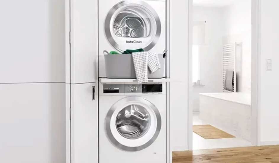 A classic white Bosch washing machine with a round front door and control dials sits on a dark, speckled base. The drum is empty and clean.