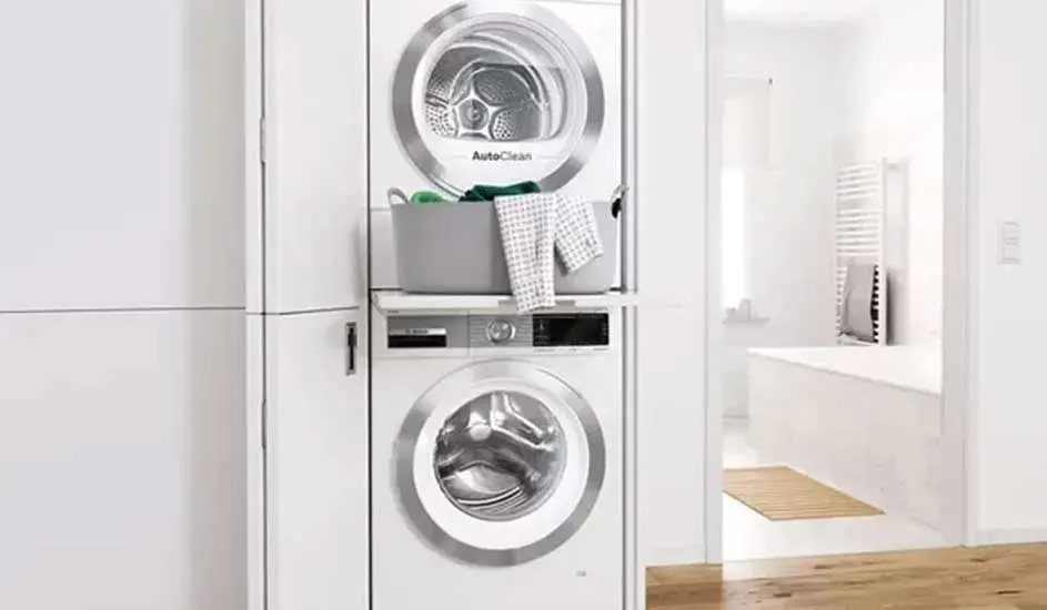 A classic white Bosch washing machine with a round front door and control dials sits on a dark, speckled base. The drum is empty and clean.