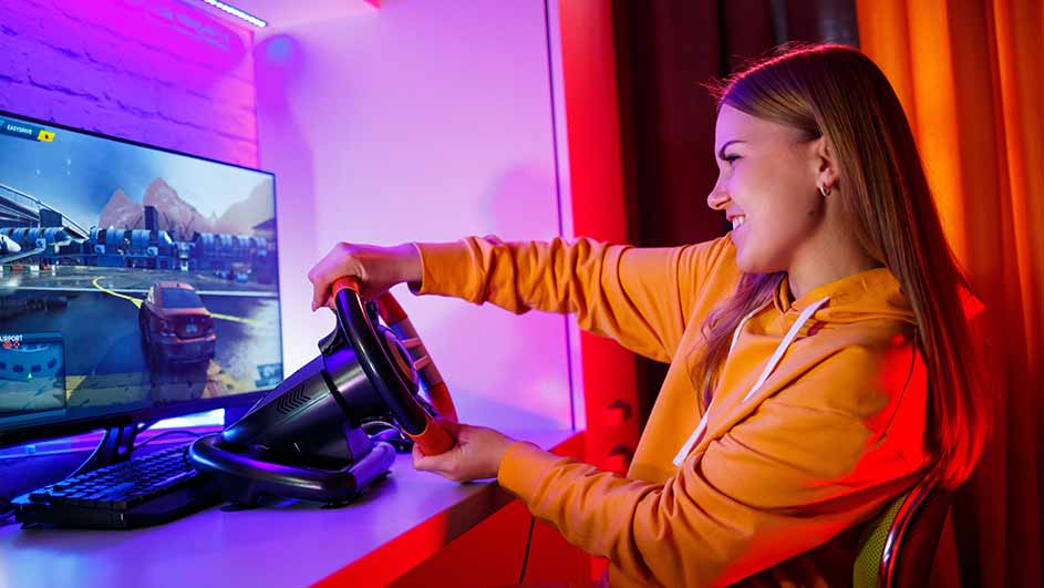 Een vrouw met een oranje hoodie speelt een racegame met een stuurwiel voor een monitor.