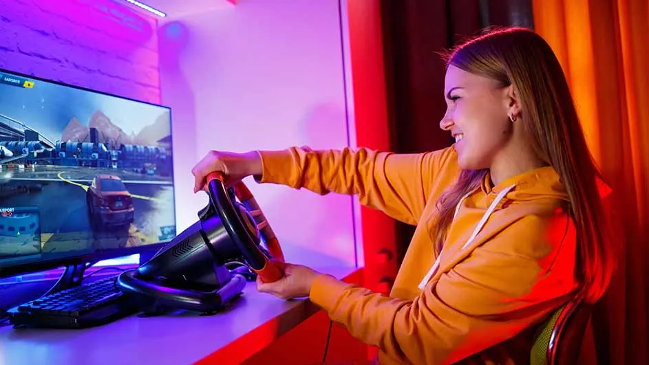 Een vrouw met een oranje hoodie speelt een racegame met een stuurwiel voor een monitor.