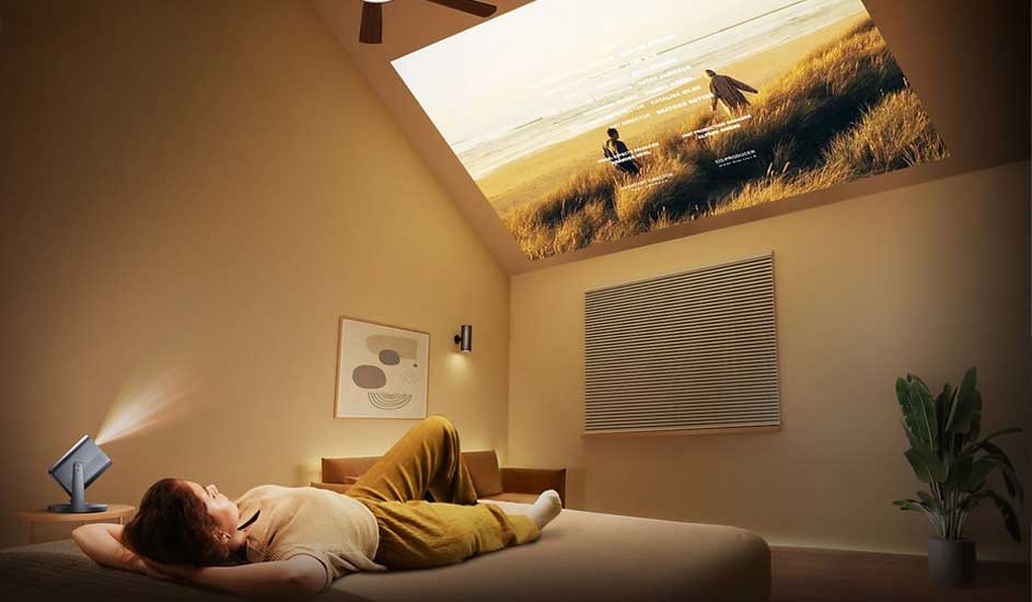 A person is lying on a bed in a cosy room, watching a film projected onto the ceiling by a Samsung portable projector. The scene shows a sunset beach.