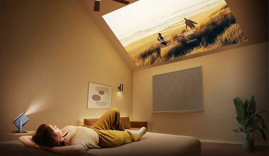 A person is lying on a bed in a cosy room, watching a film projected onto the ceiling by a Samsung portable projector. The scene shows a sunset beach.