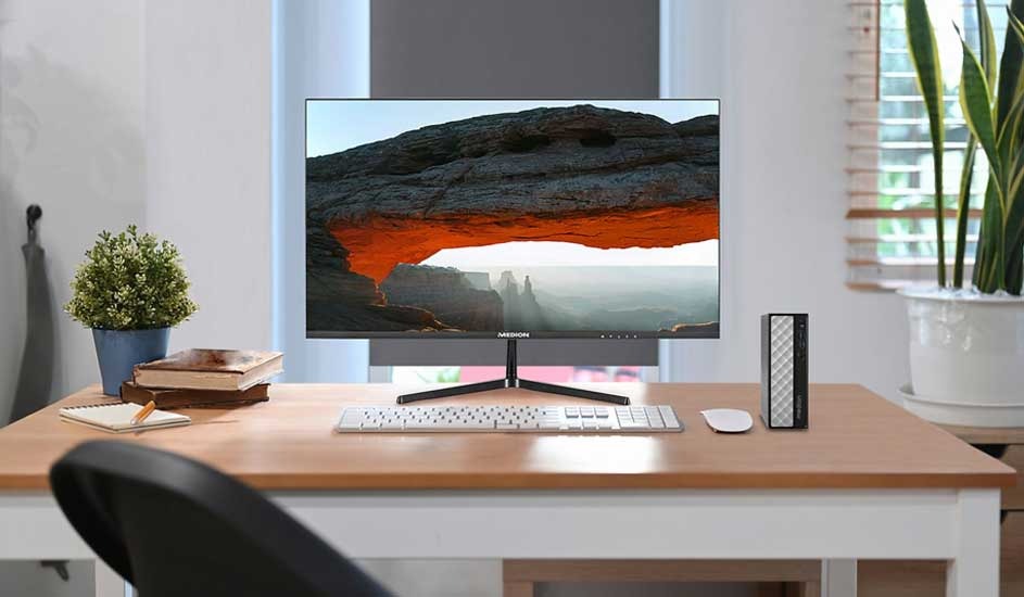Neat desk with slim monitor showing a rocky arch landscape, white keyboard and mouse, plant and books in soft daylight.