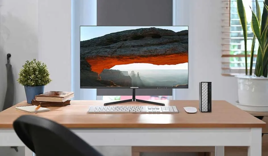Neat desk with slim monitor showing a rocky arch landscape, white keyboard and mouse, plant and books in soft daylight.