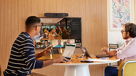Twee collega’s zitten rond een tafel en nemen deel aan een grote videogesprekssessie op een wandgemonteerd scherm.