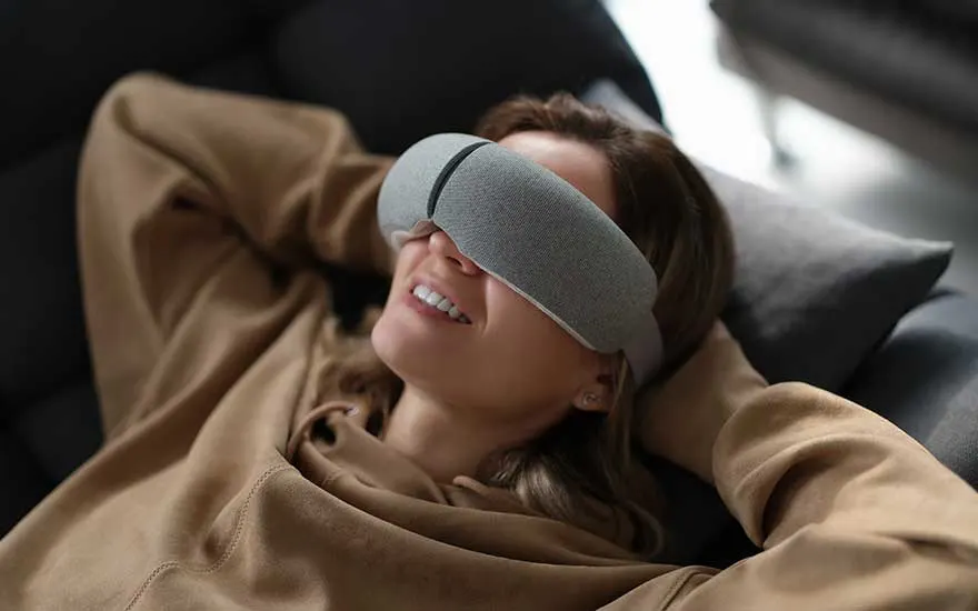 A woman relaxes on the sofa with a grey relaxation mask on her eyes, smiling and enjoying a peaceful moment at home.