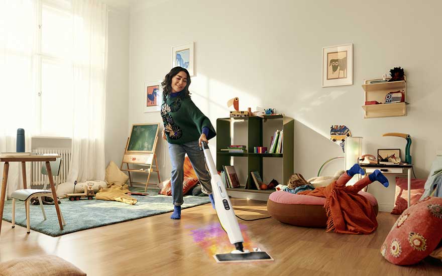 Woman steams a wooden floor in a cosy living room, while a child lounges on a cushion in the background amid toys and colourful decor.