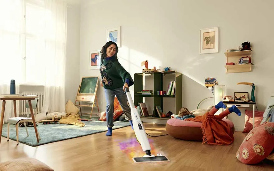 Woman steams a wooden floor in a cosy living room, while a child lounges on a cushion in the background amid toys and colourful decor.