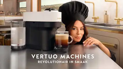 Een zwarte koffiemachine naast een glas koffie en een vrouw met een koksmuts in een keuken.