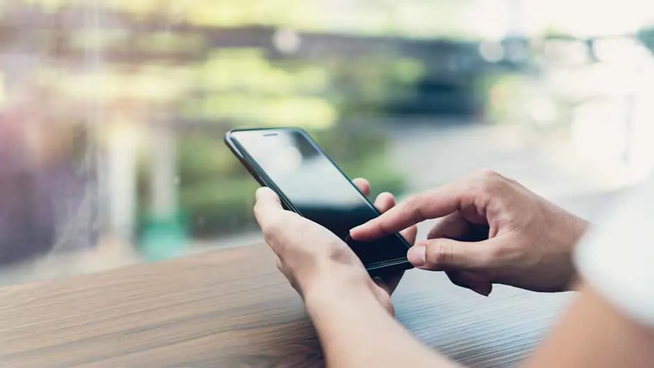 Een persoon gebruikt een zwarte smartphone op een houten tafel, met een wazige groene achtergrond.