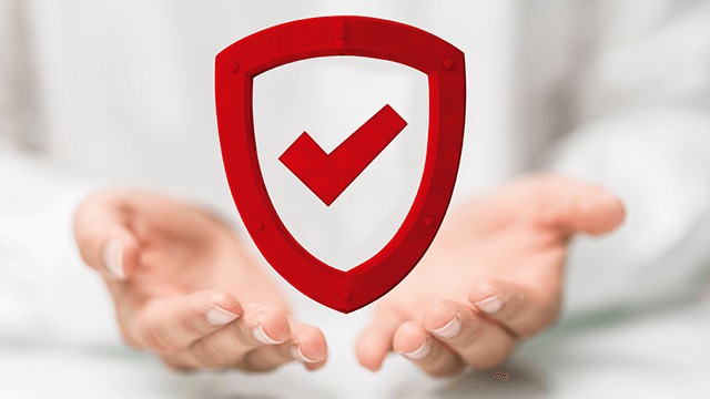 Hands hold a red shield with a checkmark. The background is blurred and white.
