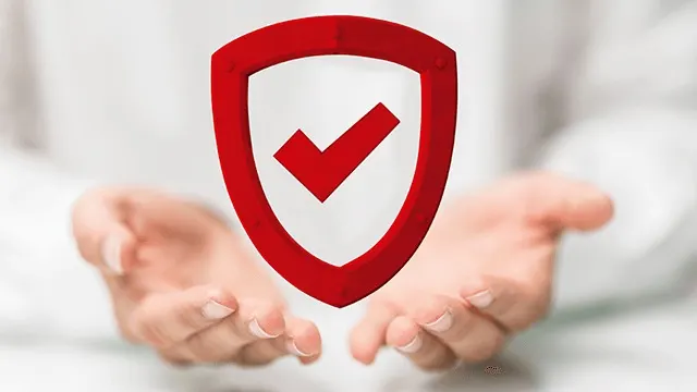 Hands hold a red shield with a checkmark. The background is blurred and white.