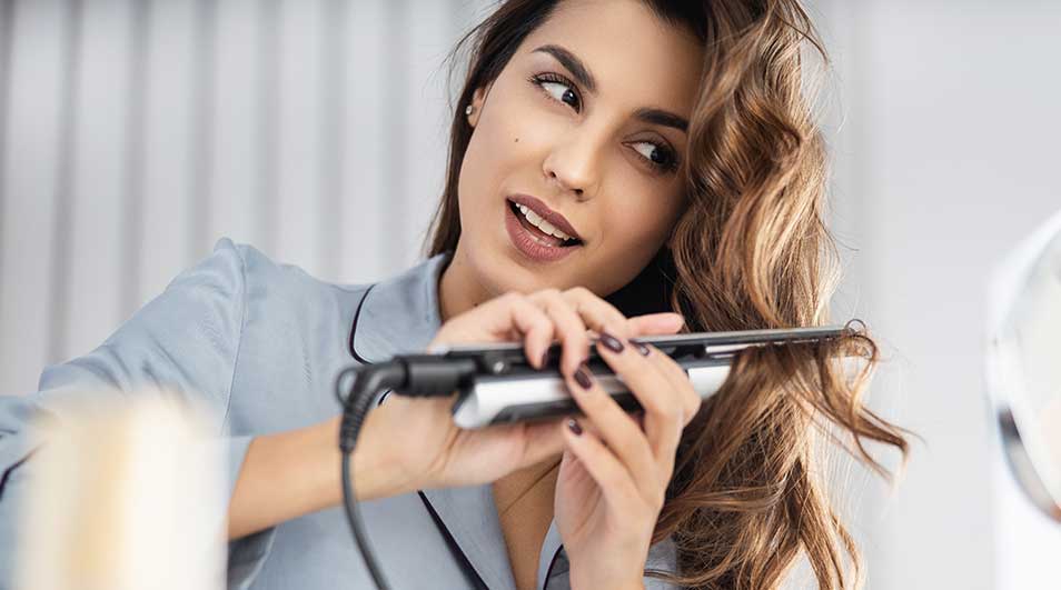 Een vrouw stylet haar golvende haar met een stijltang voor een spiegel, terwijl make-upkwasten zichtbaar zijn op de voorgrond.