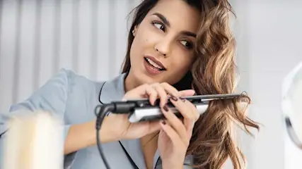 Een vrouw stylet haar golvende haar met een stijltang voor een spiegel, terwijl make-upkwasten zichtbaar zijn op de voorgrond.