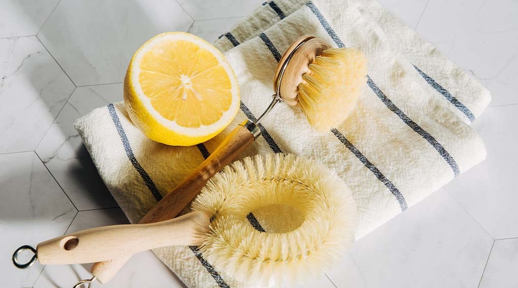 A halved lemon, two wooden dish brushes, and a striped towel are on a white, tiled surface.