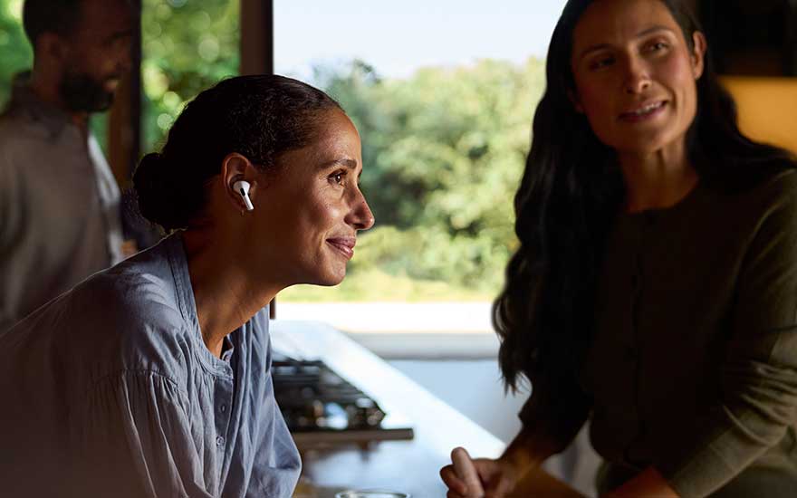 Twee mensen praten ontspannen in een lichte keuken, terwijl een vrouw met draadloze oordopjes naar muziek of een gesprek luistert.