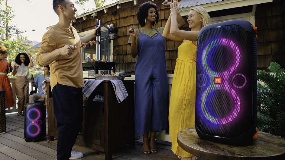 Mensen vieren feest buiten met een speaker met lichteffecten. Er staat een barbecue op tafel.