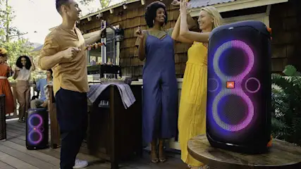 Mensen vieren feest buiten met een speaker met lichteffecten. Er staat een barbecue op tafel.