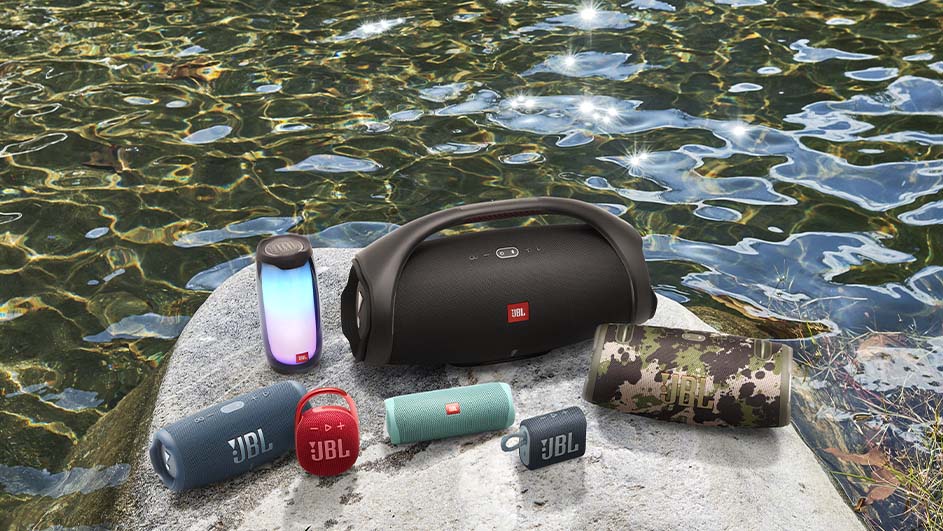 Diverse JBL-speakers staan op een rots naast water. Kleuren omvatten blauw, rood, groen, zwart en camouflage.