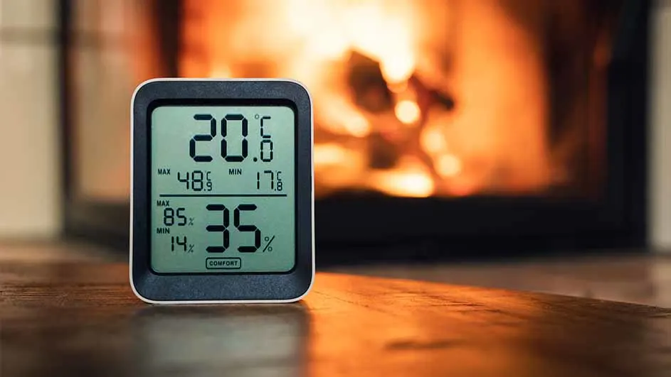 Een digitale thermometer toont 20 graden Celsius en 35% luchtvochtigheid, op een houten oppervlak voor een brandende open haard.