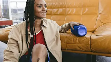 Een persoon zit op een gele leren bank met een blauwe Fresh 'n Rebel Bluetooth speaker in de hand. De sfeer is ontspannen en casual.