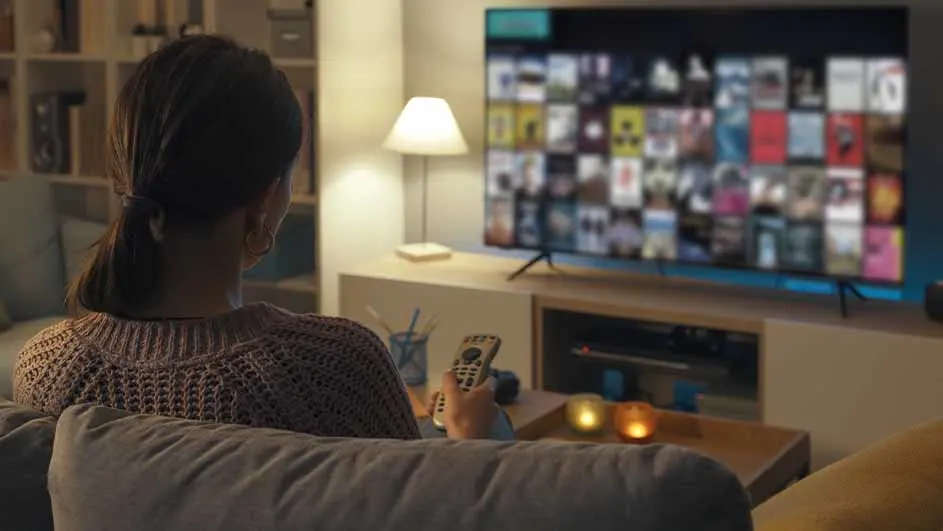 Vrouw zit op een bank met een afstandsbediening in haar hand, kijkend naar een tv in een woonkamer.