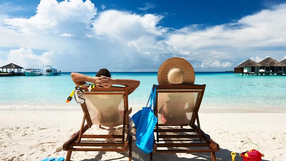 Twee ligstoelen op een strand met zicht op een blauwe zee, een boot en huizen op palen onder een blauwe lucht met witte wolken.