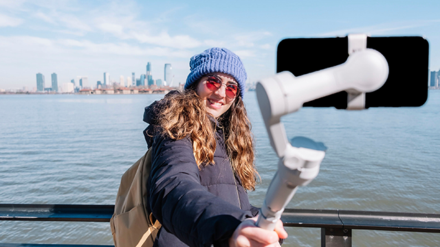 Een persoon met een blauwe muts en zonnebril houdt een telefoon aan een stabilisator vast. Op de achtergrond is water en een skyline.