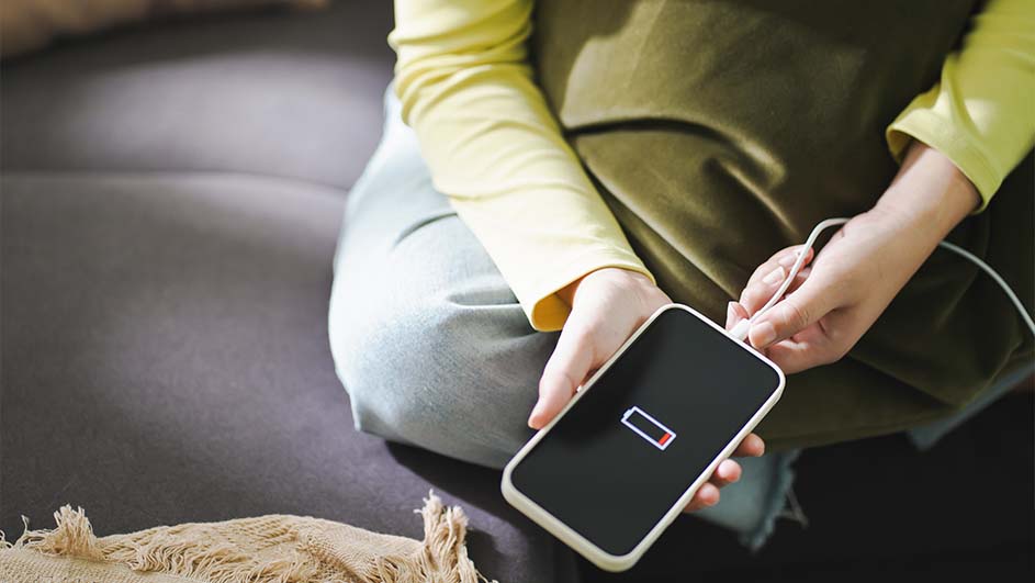 Zittend persoon met geel shirt en groene broek sluit witte kabel aan op een zwarte telefoon met lege batterij op het scherm.