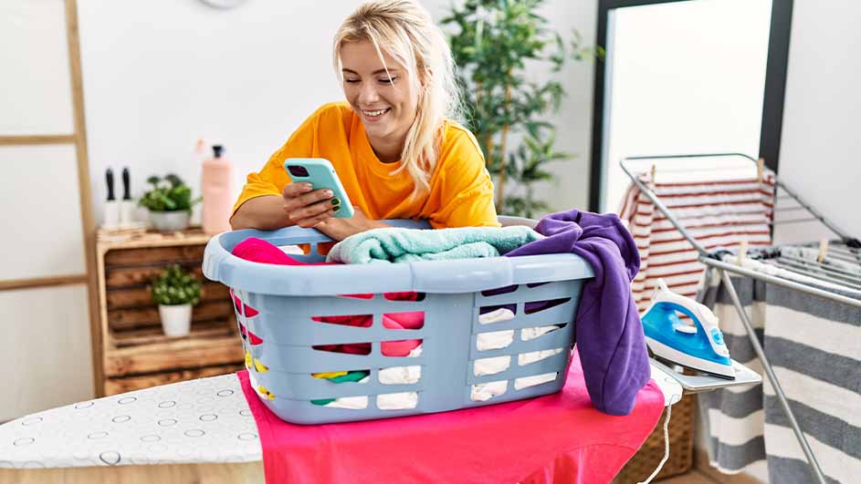 Vrouw leunt op wasmand met kleurrijke was, met telefoon in de hand. Strijkplank, droogrek en planten zichtbaar.