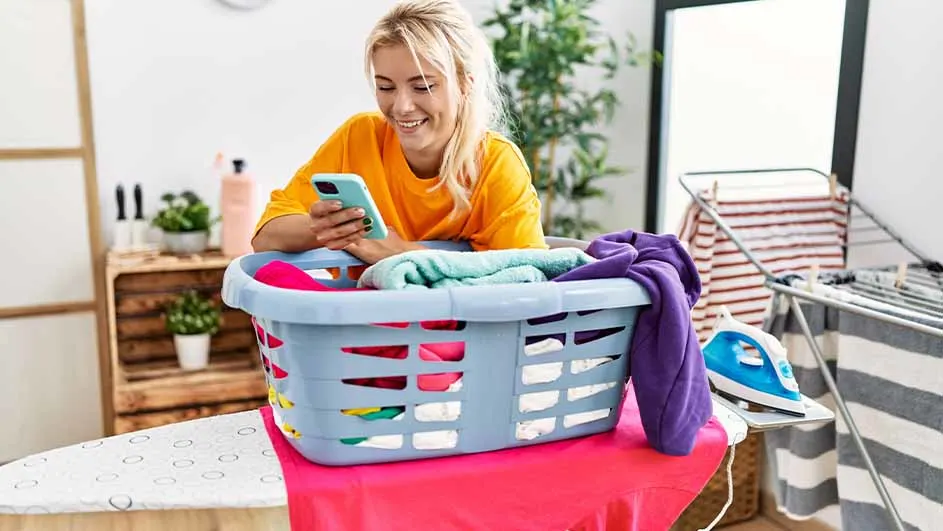 Vrouw leunt op wasmand met kleurrijke was, met telefoon in de hand. Strijkplank, droogrek en planten zichtbaar.