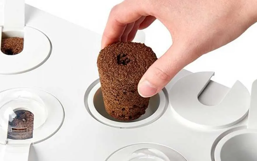 Een hand plaatst een bruine groeipod in een witte smart garden. Het apparaat heeft meerdere openingen voor plantencapsules.