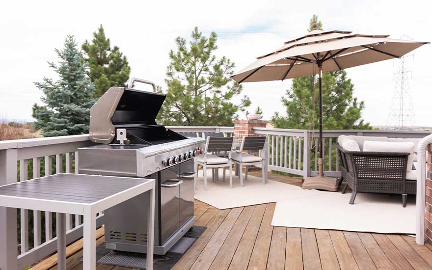 Een ruim houten terras toont een moderne barbecue, tuinmeubels en een grote parasol in een rustige buitenomgeving.