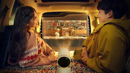 Twee mensen in een auto kijken naar een projectie van een platenzaak, een witte projector staat op een kleurrijk kleed.