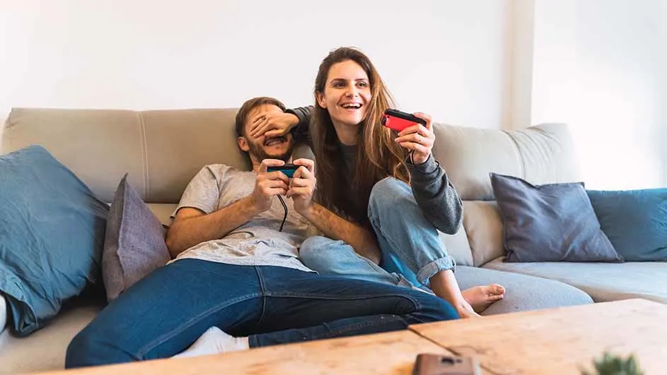 Een vrouw en een man zitten op een bank met blauwe kussens, gamen met rode en blauwe controllers.