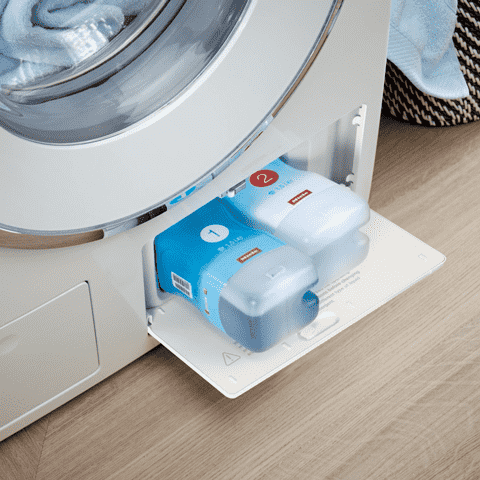 Wasmachine met blauwe en witte flessen wasmiddel in een lade. Er ligt wasgoed in de trommel.