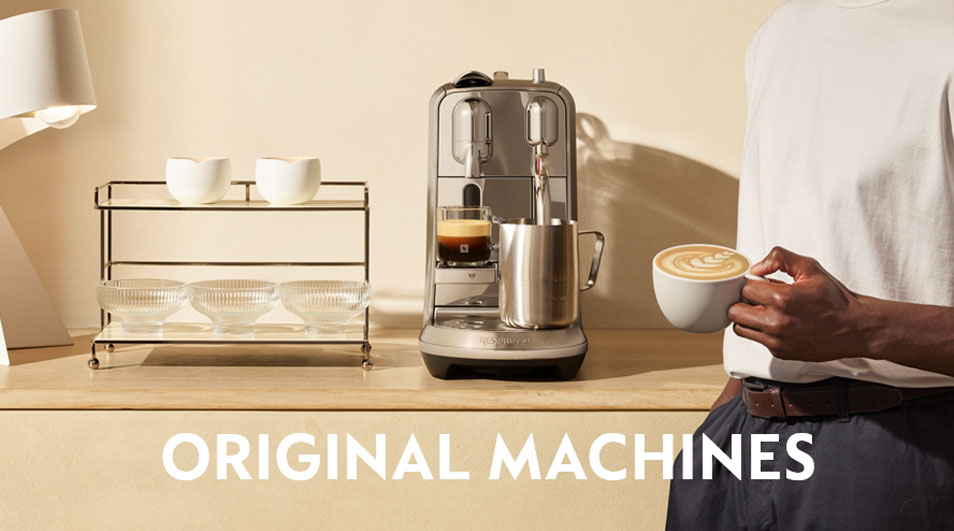 Koffiehoek met een Nespresso Original-machine op een houten blad; een persoon houdt een cappuccino vast, met lamp en rek met kopjes op de achtergrond.