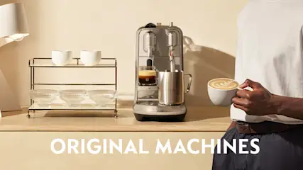 Koffiehoek met een Nespresso Original-machine op een houten blad; een persoon houdt een cappuccino vast, met lamp en rek met kopjes op de achtergrond.