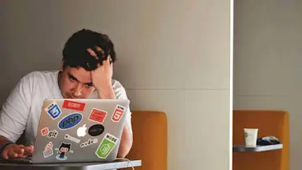 Een man zit achter een laptop met stickers, hand aan het hoofd, met een beker en zonnebril op een plank in de achtergrond.