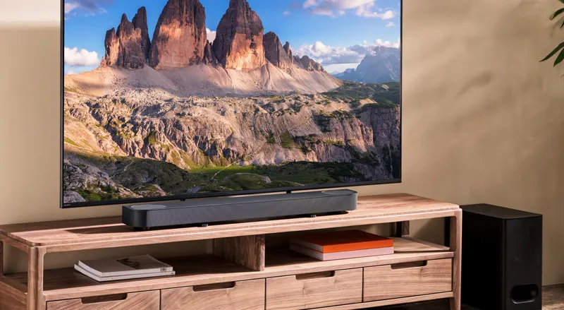 Strak houten tv-meubel met centrale zwarte soundbar en subwoofer ernaast, bovenop het meubel een flatscreen-tv die een berglandschap toont, in een minimalistisch interieur.