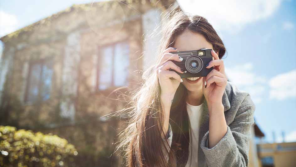 Een vrouw glimlacht en houdt een zwarte camera voor haar gezicht. Op de achtergrond staan een gebouw en een blauwe lucht.