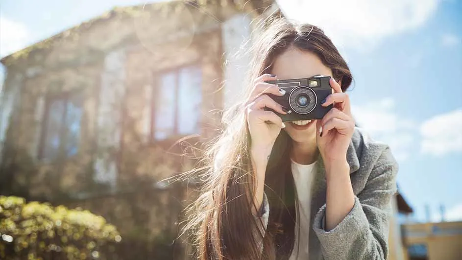 Een vrouw glimlacht en houdt een zwarte camera voor haar gezicht. Op de achtergrond staan een gebouw en een blauwe lucht.